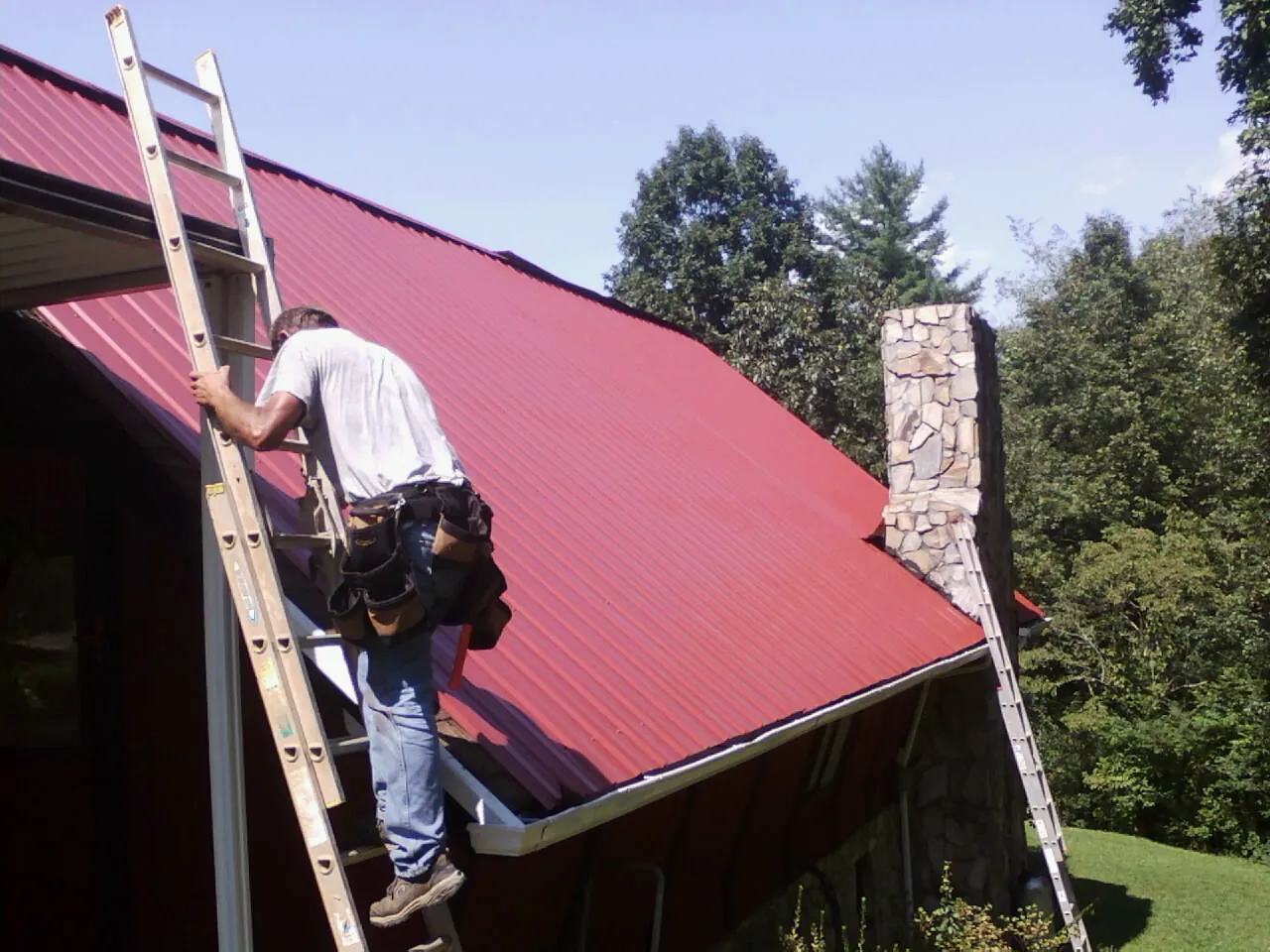 Roofer on a ladder beside a red metal roof and stone chimney — Soffit & Fascia Repair by Roofing Brothers in Owensville